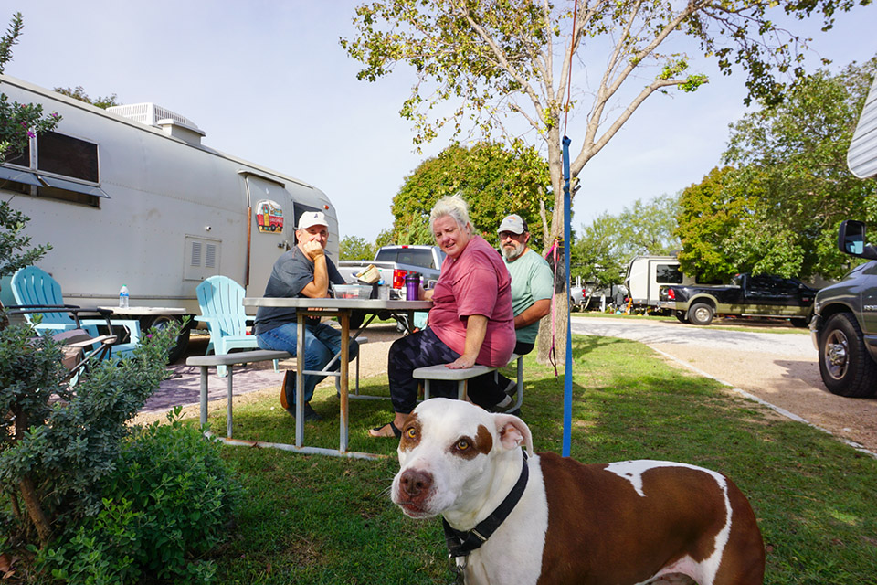 Three people sitting at an outdoor table next to a silver Airstream trailer in an RV park