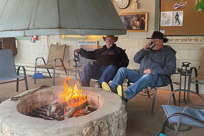 Two men relaxing in front of an indoor fire pit, each holding a drink and wearing casual outdoor clothing and cowboy hats