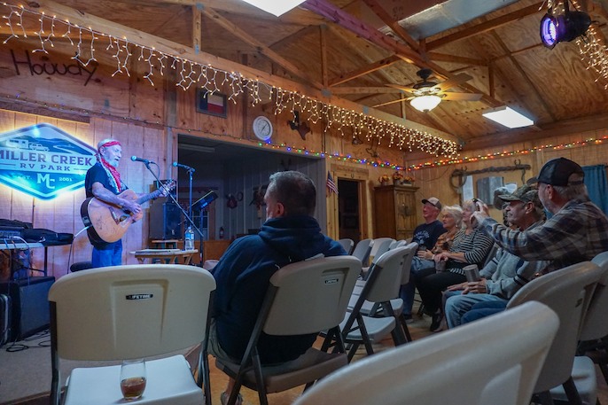 Small audience seated in an indoor hall enjoying a live music performance, with the performer playing an acoustic guitar on a stage decorated with a Miller Creek RV Park sign and string lights