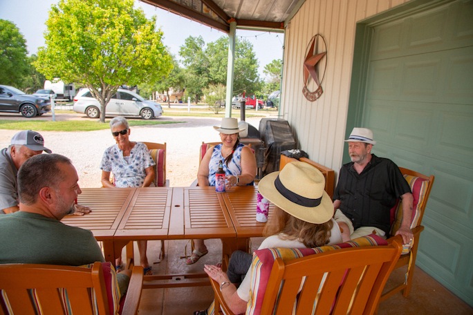 Group of people seated around a wooden table on a covered porch, enjoying a sunny day at an RV park