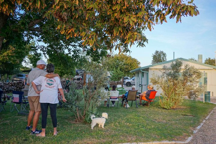 people sitting in a semi-circle near a small building, a couple with a small white dog in the foreground, and greenery along with RVs in the background