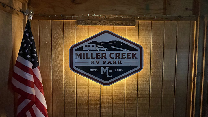 Illuminated Miller Creek RV Park sign on a wooden wall with an American flag on the left