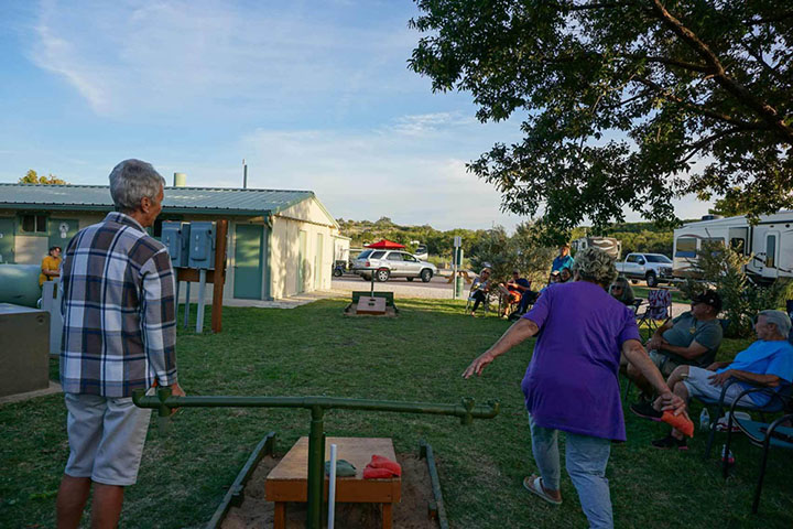 Retired RV park residents enjoying an outdoor game of cornhole, with an older man and woman playing in the foreground and other residents watching in the background