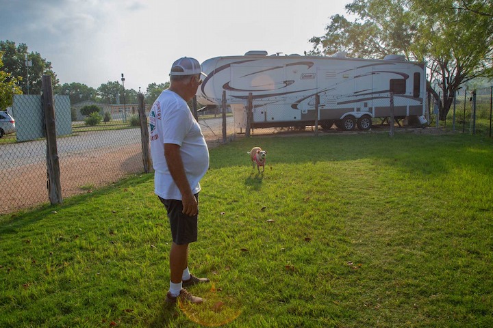 Man standing inside a fenced dog run area at an RV park, looking at a small dog running towards him, with a large RV and trees in the background