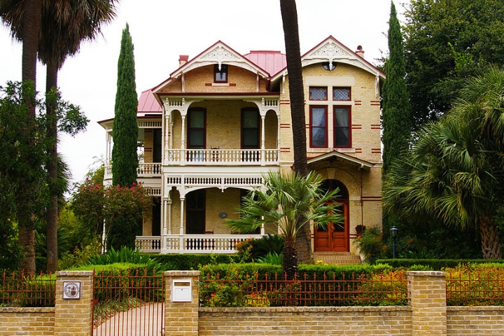 A beautifully preserved Victorian-style house in the King William Historic District, featuring intricate wooden detailing, tall windows, and lush landscaping surrounded by a brick fence