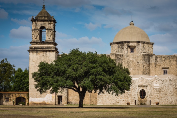 The historic Mission San José in San Antonio, showcasing its distinctive dome, bell tower, and stone walls, with a prominent tree in front