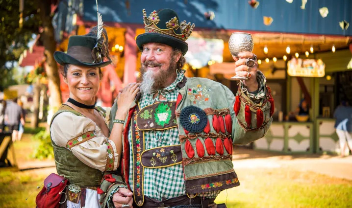 A jovial couple dressed in colorful Renaissance-era costumes, with the man raising a goblet at the Texas Renaissance Festival