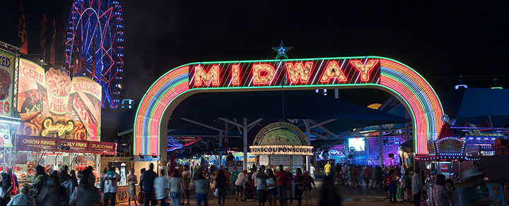 The illuminated 'Midway' arch at night, welcoming visitors to the Texas State Fair with food vendors and carnival rides in the background