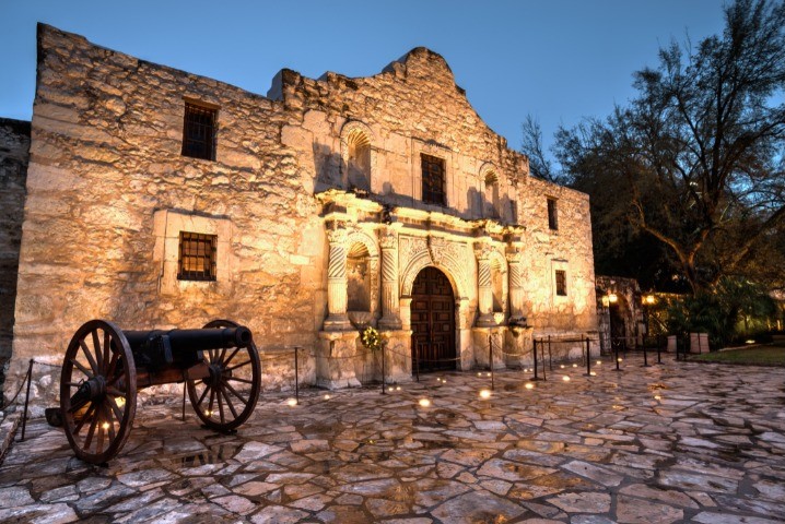 The historic stone facade of The Alamo in San Antonio, Texas, illuminated at dusk, with a cannon displayed in the foreground on the cobblestone plaza