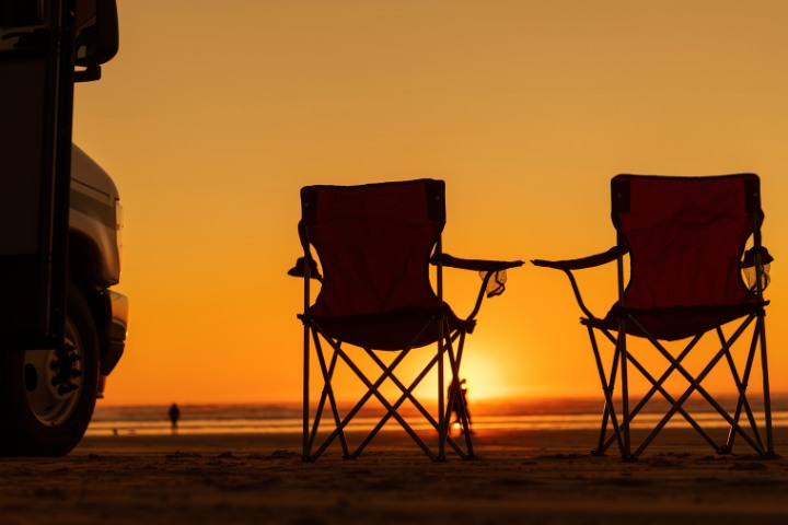 Two camping chairs set up near an RV, facing a sunset at the beach