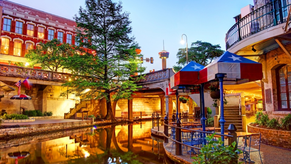 The lively San Antonio River Walk, featuring a tree-lined canal, outdoor seating with Texas flags, and historic buildings illuminated in the evening light