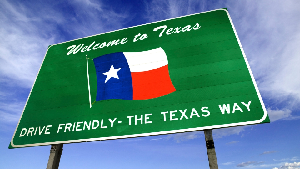 A green highway sign reading 'Welcome to Texas' with the Texas flag and the phrase 'Drive Friendly - The Texas Way' beneath it, set against a bright blue sky