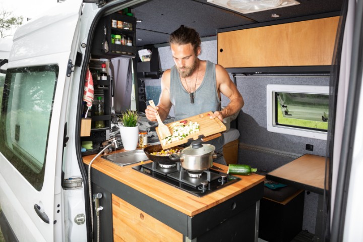 Camper cooking in a well-equipped RV van kitchen, using a cutting board and stove for meal preparation