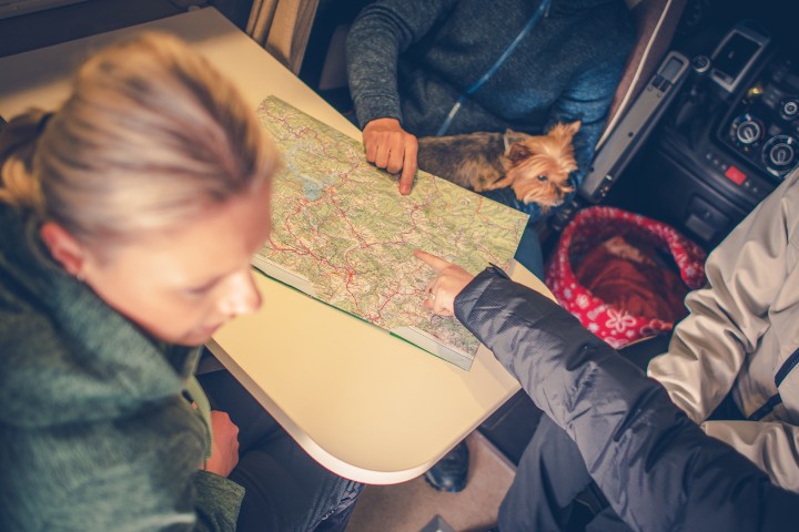 A group of travelers inside an RV planning their route using a map, with a small dog nearby