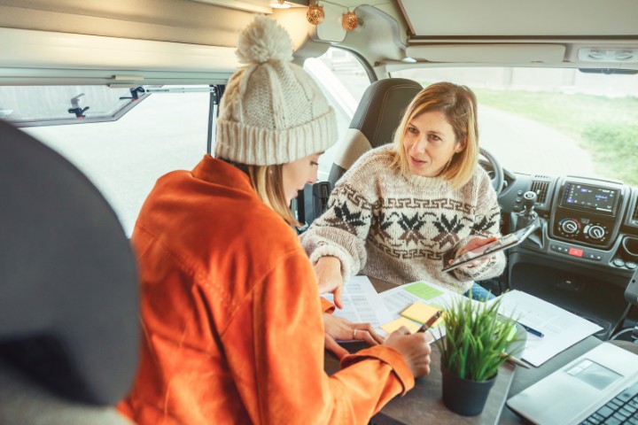 Two women inside an RV planning their camping trip, with maps and notes spread across the table