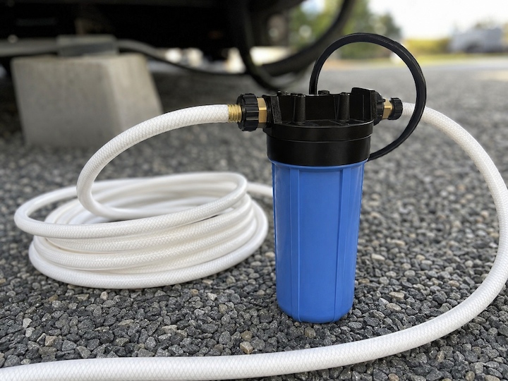 Blue inline RV water filter connected to a coiled white drinking water hose on gravel.