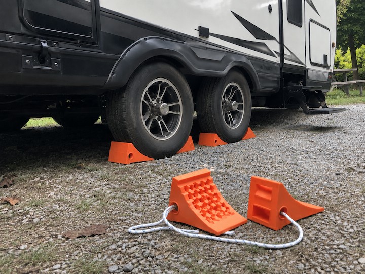Orange wheel chocks securing both sides of travel trailer wheels at a gravel RV campsite