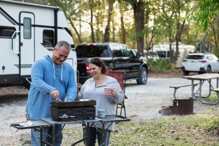 Couple cooking outside their RV at a campsite with a tow vehicle and RV parked nearby