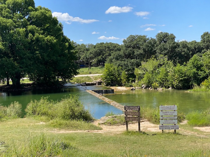 Natural river swimming area and dam at Blanco State Park with park safety signs along the shore